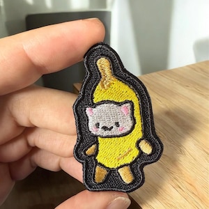 May include: Embroidered patch featuring a cartoon cat dressed as a banana. The cat has a gray face with pink ears and a small smile. The banana is yellow with brown accents, and the patch has a black border.