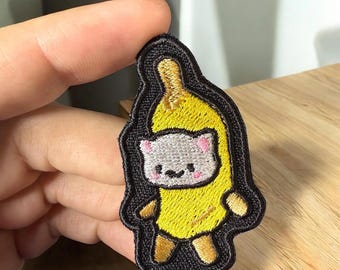 May include: Embroidered patch featuring a cartoon cat dressed as a banana. The cat has a gray face with pink ears and a small smile. The banana is yellow with brown accents, and the patch has a black border.