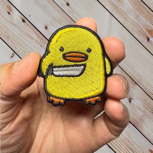 May include: Embroidered yellow chick patch with a black outline, holding a silver knife. The chick has an orange beak and feet. The patch is held in a hand against a light wood grain background.