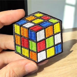 May include: Embroidered patch featuring a colorful Rubik's Cube design. The cube is composed of squares in various colors, including red, orange, yellow, green, blue, and white, outlined in black. The patch is held against a light wooden surface.