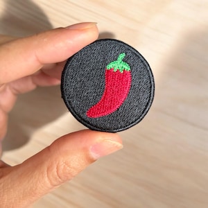 May include: A round, black embroidered patch featuring a red chili pepper with a green stem. The patch is held up by a hand. The chili pepper is the focal point, with detailed stitching.