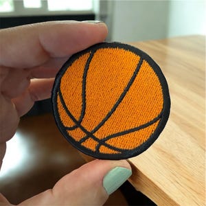 May include: Round orange basketball patch with black stitched details. The patch features the classic basketball design with black lines. The patch is held by a hand.