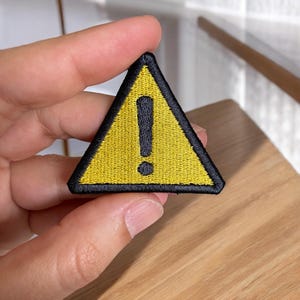 May include: Embroidered yellow triangle patch with a black border and a black exclamation point in the center. The patch is a warning symbol, commonly used to indicate caution or a potential hazard. The patch is held by a person.