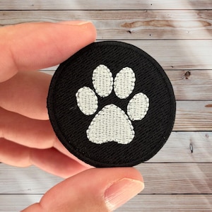 May include: A round, black patch featuring a white paw print design. The paw print is embroidered with a textured, stitched appearance. The patch is held in a person's hand against a wooden background. This patch is suitable for pet lovers.