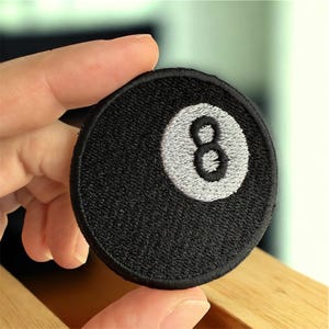 May include: A round, black patch featuring a white embroidered number 8, resembling a billiard ball. The patch has a textured surface and a stitched border. Ideal for jackets, bags, or other fabric items.