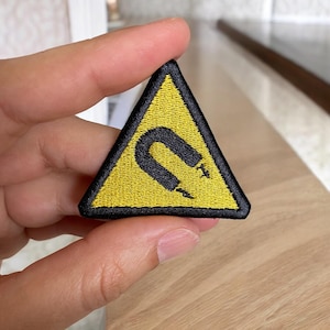 May include: Embroidered patch in a yellow triangle shape with a black border. The patch features a black horseshoe magnet symbol. The patch is held in a person's hand.