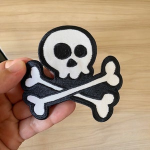 May include: White skull and crossbones patch with black stitching. The skull has black eyes and a black nose.
