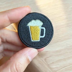 May include: A black embroidered patch with a white outline of a beer mug filled with yellow beer and a white foam head.