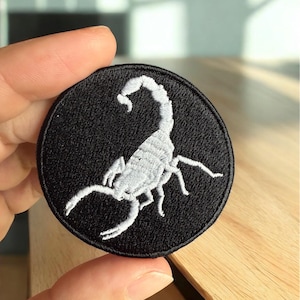 May include: A round, black embroidered patch featuring a detailed white scorpion design. The scorpion is centered on the patch, with its pincers and tail clearly visible. The patch is likely for clothing or accessories.