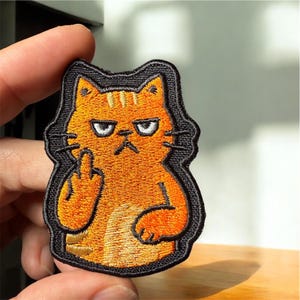 May include: Embroidered patch featuring an orange cat with an angry expression, giving the middle finger. The cat is outlined in black, with detailed stitching. A humorous and edgy accessory for clothing or bags.