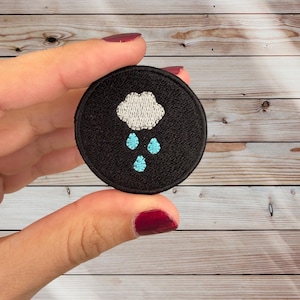 May include: A black embroidered patch with a white cloud and blue raindrops. The patch is circular and has a black border.