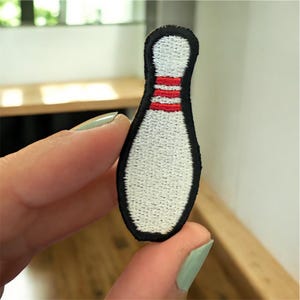 May include: Embroidered bowling pin patch with a white body, three red stripes, and a black border. The patch is held between fingers, showcasing its size and detail. This patch is ideal for adding to clothing or accessories.
