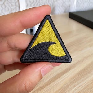 May include: A yellow triangle-shaped patch with a black wave design. The patch is embroidered with black and yellow thread.