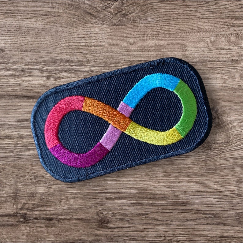 Puede incluir: Parche bordado con un s&iacute;mbolo de infinito multicolor, que representa la neurodiversidad. El s&iacute;mbolo est&aacute; compuesto por colores del arco&iacute;ris, incluyendo rosa, naranja, morado, azul y verde, cosidos sobre un fondo azul marino. Forma rectangular con esquinas redondeadas.