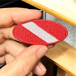 May include: Oval-shaped red and white patch with a diagonal white stripe. The patch is embroidered with a textured appearance. The design is a diving flag, commonly used to indicate a diver is in the water.