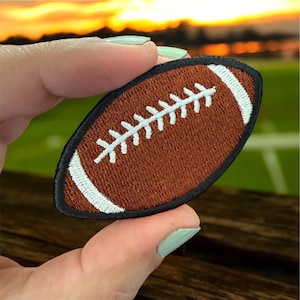 May include: Embroidered patch in the shape of a brown football with white stitching and accents. The patch has a black border. The patch is held up against a blurred background of a sunset and a football field.