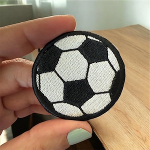 May include: Round embroidered patch featuring a soccer ball design. The patch has a black border and a black and white soccer ball pattern. The patch is held by a hand.