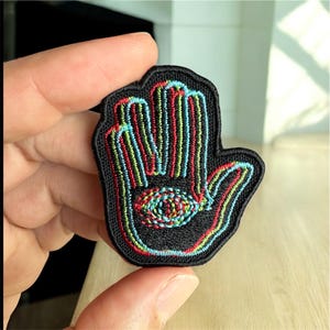 May include: Embroidered black patch in the shape of a hand with a central eye design. The patch features colorful stitching in red, blue, and green, creating a vibrant and detailed design. The patch is likely for clothing or accessories.