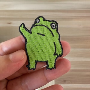 May include: Green embroidered frog patch with black outline and white eyes. The frog is standing on its hind legs with its arms raised in the air.