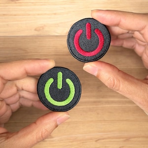 May include: Two round black fabric patches with embroidered power button symbols. One patch has a red power button symbol and the other has a green power button symbol.