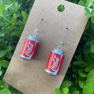 May include: A pair of silver earrings with red Coca-Cola cans hanging from them. The cans are silver with red lettering and a white logo.