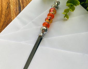 May include: A decorative letter opener with a silver blade and handle. The handle features orange and yellow beads with silver accents. The opener is resting on white paper, with greenery in the background.