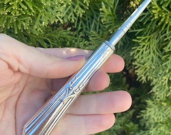 May include: A silver-toned antique ear spoon with a decorative handle and a small scoop at the end. The handle features an engraved floral design and vertical lines. The text "E/4" is visible on the shaft.