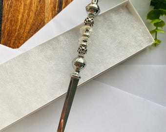 May include: A silver letter opener with a decorative handle featuring clear and silver beads. The opener is displayed in a white box, with a wooden background and green foliage in the corner.