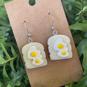 May include: A pair of silver dangle earrings featuring a white ceramic toast with two yellow fried eggs on top.