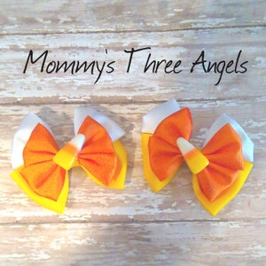 May include: Two orange, yellow, and white candy corn hair bows with white ribbon accents.  "Mommy's Three Angels" is written in black cursive above the bows.