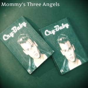 May include: Two vintage "Cry-Baby" movie promotional cards featuring a portrait of the actor. The cards have a dark green background with the movie title in white script. The cards are slightly angled, with the actor's image visible.