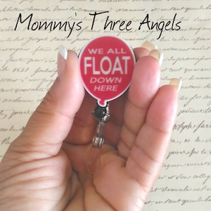 May include: A red balloon with the text "WE ALL FLOAT DOWN HERE" on it. The balloon is attached to a black and silver retractable badge holder.