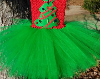 May include: A green tulle tutu with a red crocheted bodice featuring a green felt Christmas tree with a gold star and colorful lights. The text "Mommy's Three Angels" is visible at the bottom of the image.