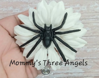 May include: A white flower with a black spider on top. The flower is attached to a clear plastic badge holder. The text "Mommy's Three Angels" is visible below the flower.