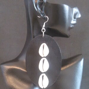May include: A pair of large, round, black earrings with three white cowrie shells hanging from them. The earrings are on a black mannequin head.