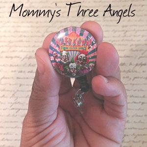 May include: A round, retractable badge reel featuring the text "Killer Klowns From Outer Space" with images of scary clowns. The reel is held in a hand, and the text "Mommy's Three Angels" is above the reel.