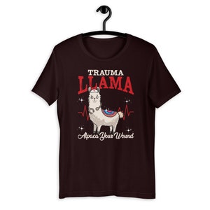 K&ouml;nnte beinhalten: Ein dunkelbraunes T-Shirt mit einem Cartoon-Lama, das eine Krankenschwesterm&uuml;tze und ein Stethoskop tr&auml;gt. Der Text "Trauma Llama" befindet sich &uuml;ber dem Lama und "Alpaca Your Wound" unter dem Lama.