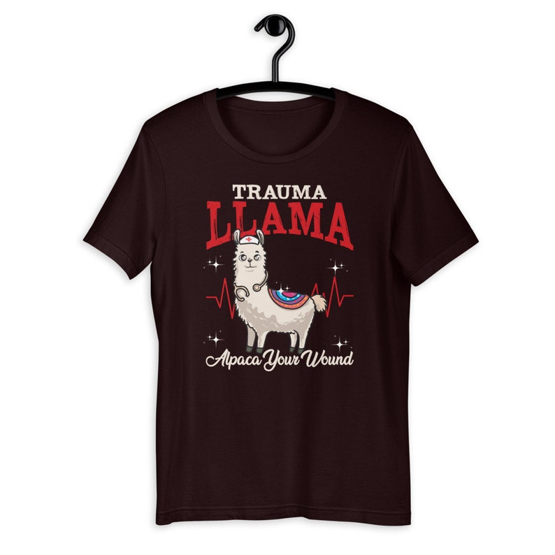 K&ouml;nnte beinhalten: Ein dunkelbraunes T-Shirt mit einem Cartoon-Lama, das eine Krankenschwesterm&uuml;tze und ein Stethoskop tr&auml;gt. Der Text "Trauma Llama" befindet sich &uuml;ber dem Lama und "Alpaca Your Wound" unter dem Lama.