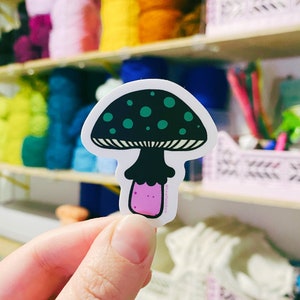 The artist holds up a hand drawn vinyl sticker in the shape of a funky toadstool mushroom, in her art studio.