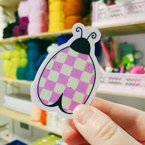 The artist holds a hand drawn vinyl sticker in the shape of a beetle with a purple checkerboard pattern, in her art studio.