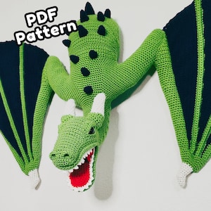 May include: A crochet dragon wall hanging featuring a bright green body, dark navy blue wings with green accents, white horns, and an open mouth with a red interior. The text PDF Pattern is visible in the upper left corner of the image.