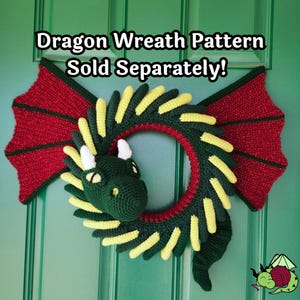 K&ouml;nnte beinhalten: Ein geh&auml;kelter Drachenkranz mit roten Fl&uuml;geln und einem gr&uuml;nen Kopf. Der Kranz ist gr&uuml;n mit gelben Akzenten und einem roten Innenring. Der Text auf dem Bild lautet "Dragon Wreath Pattern Sold Separately!"