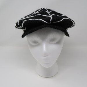 May include: A black and white knitted newsboy cap with a white spiderweb design.