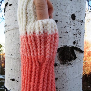 May include: A pair of knitted arm warmers with a white top and a coral pink bottom. The arm warmers are made with a textured stitch.