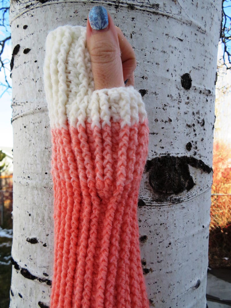 May include: A pair of knitted arm warmers with a white top and a coral pink bottom. The arm warmers are made with a textured stitch.