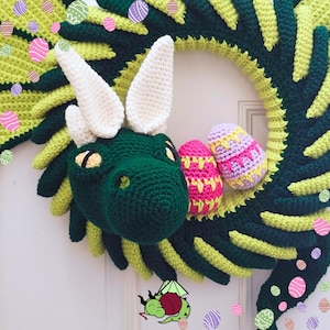 K&ouml;nnte beinhalten: Ein handgefertigter H&auml;kelkranz mit einem gr&uuml;nen Drachenkopf mit wei&szlig;en H&ouml;rnern und Ohren sowie gelben Augen. Der Kranz ist mit rosa und lila geh&auml;kelten Ostereiern verziert. Der Kranz ist gr&uuml;n und gelb.