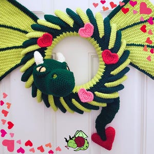 K&ouml;nnte beinhalten: Ein handgefertigter H&auml;keldrachen-Kranz in Gr&uuml;n- und Gelbt&ouml;nen, verziert mit roten und rosa Herzen. Der Drache hat einen detaillierten Kopf mit wei&szlig;en H&ouml;rnern und gelben Augen. Der Kranz ist zum Aufh&auml;ngen an einer T&uuml;r gedacht.