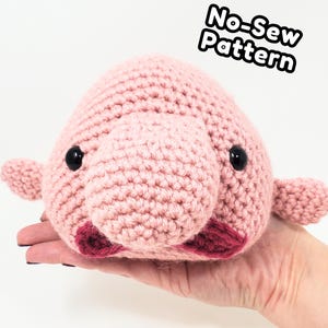 Może przedstawiać: Szydełkowana różowa pluszowa ryba-blob z czarnymi oczami i bordowymi ustami. Pluszak jest trzymany w dłoni i ma małe płetwy. Tekst "No-Sew Pattern" znajduje się w prawym górnym rogu.
