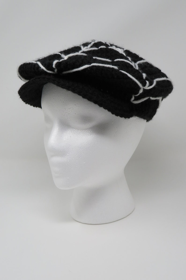 May include: A black and white patterned crocheted newsboy cap. The cap has a white brim and a black crown with a white geometric design.