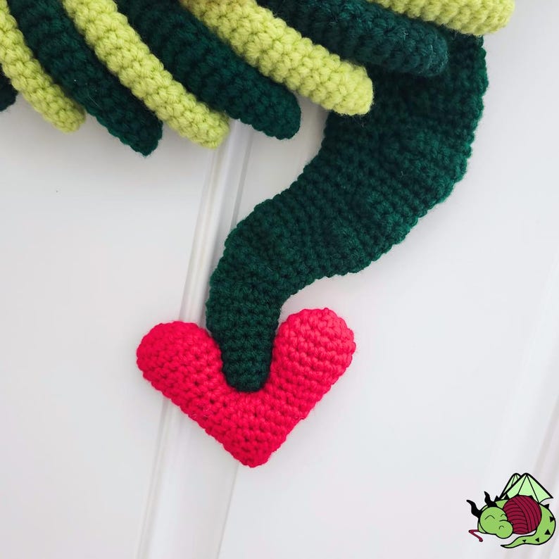 K&ouml;nnte beinhalten: Geh&auml;kelter gr&uuml;ner und gelber Kranz mit rotem Herzakzent. Der Kranz besteht aus dickem Garn und hat eine strukturierte Optik. Das Herz ist leuchtend rot und am unteren Ende des Kranzes befestigt.